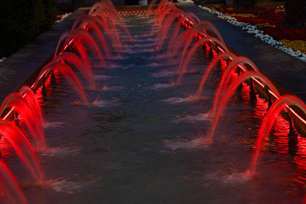 Red-lit water fountains in a garden setting, creating a vibrant night scene with reflections on the water.