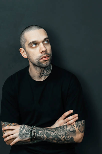 Tattooed man wearing a black shirt looks upwards with a thoughtful expression. Visible tattoos on arms and neck, suggesting contemplation or deep thought against a dark background. High quality photo