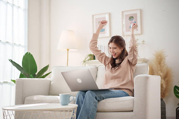istock A joyful woman raises her arms in celebration while sitting on a comfortable couch, working on her laptop in a well-lit living room. She enjoys her productive moment 2210701843