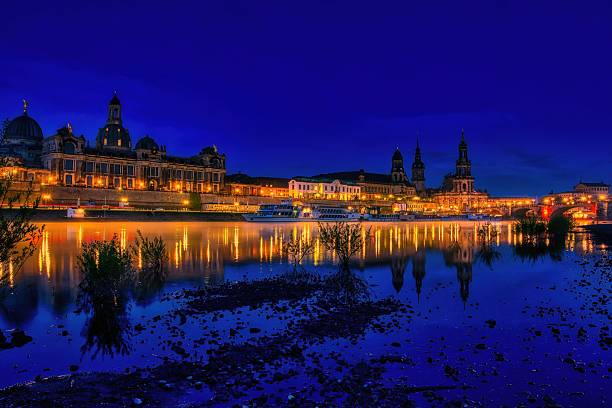 Night view of Dresden's illuminated historic buildings reflecting in the Elbe River under a deep blue sky.