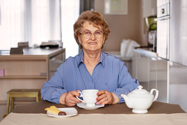 istock Happy Senior woman enjoying tea at home, smiling while holding cup, sitting at kitchen table with teapot and sweets in cozy and modern indoor 2210705164