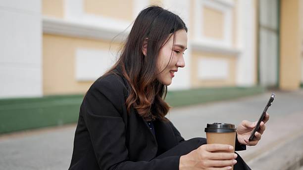 istock young asian woman sitting outdoors holding coffee and using smartphone with gentle smile 2210706162