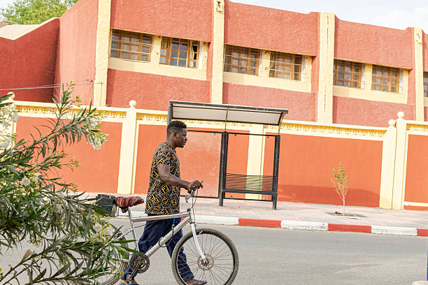 Tamanrasset, Algeria- March 22, 2025:  while loading a car battery on the rear rack, in front of a red and yellow building with a bus stop;