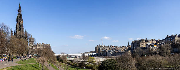 A wide panoramic shot of East Princess Street Gardens in Edinburgh. Unrecognisable people are walking and sitting around the area while enjoying the sunny weather. Landmarks such as the Scott Monument can be seen.\n\nVideos are available similar to this scenario.
