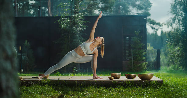 istock Serene Caucasian Female Practices Yoga in a Tranquil Forest Setting, Gracefully Transitioning Through Poses on a Wooden Platform with an Incense Stick, Surrounded by Nature and Singing Bowls 2210735731