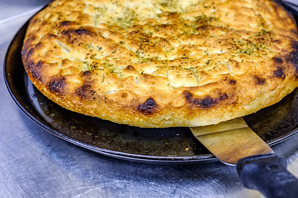A large, round flatbread with a crispy, golden crust sits on a metal counter, showing its texture and fragrant seasonings, ready for slicing and serving.