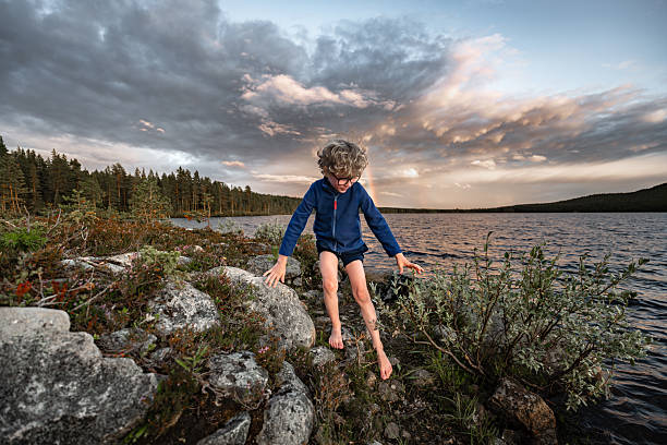 A young boy plays barefoot on the rocky shore of a serene lake in Sweden, enjoying the beauty of summer and the tranquility of the evening sky filled with clouds