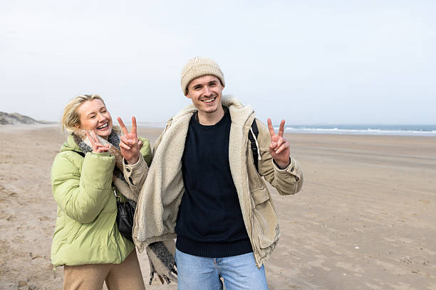 Three-quarter length front-view of a young man and woman dressed in warm, casual outdoor clothing, enjoying a day out in Newton-by-the-Sea, Alnwick. They are walking along a scenic British beach while smiling and having fun. They are making peace sign gestures.\n\nVideos are available similar to this scenario.