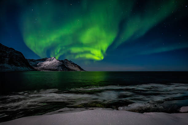 A sweeping arc of green aurora borealis lights up the night sky and reflects over the icy waters of Senja Island’s rugged coastline. Snow-covered rocks and gentle waves frame this stunning Arctic winter scene under a canopy of stars.