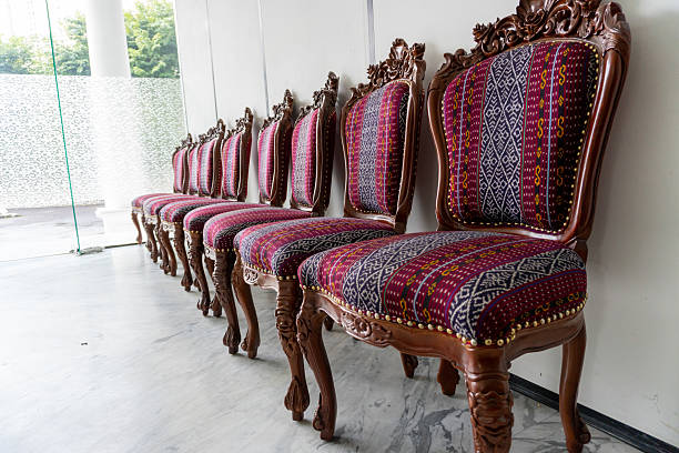 This image highlights a beautifully preserved antique armchair located in the Ambronay town hall, in the mayor's office. The chair, upholstered in rich green velvet, features intricately carved wooden arms and a high back, exuding elegance and historical charm. The backdrop of the image showcases a wall adorned with luxurious red and gold damask wallpaper, complementing the detailed woodwork and stained glass window. Photo taken in Ambronay, in Ain department, in Auvergne-Rhone-Alpes region in France.