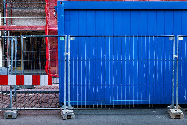 A striking urban scene of a Berlin construction site, featuring a vibrant blue metal container, red scaffolding mesh on the building facade, and a temporary safety fence with a warning light. The colors create a bold and graphic composition symbolizing city renovation and street-level infrastructure upgrades.