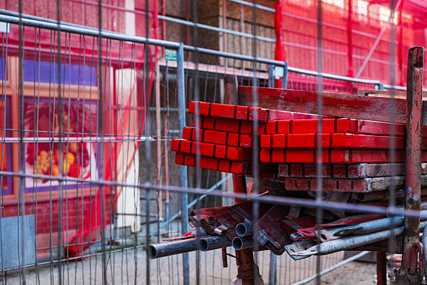 A neatly stacked bundle of red and silver metal scaffolding pipes and frames lies on a cobblestone pavement inside a fenced-off construction zone in Berlin. The image captures construction site equipment in detail, emphasizing infrastructure and urban development elements.