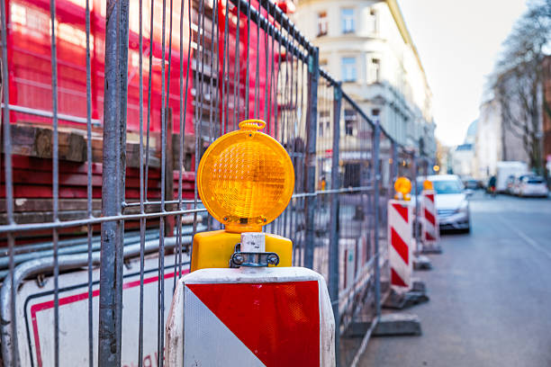 Close-up view of a yellow flashing warning light attached to a temporary road sign and metal fence in front of a blue container at a Berlin construction site. The image captures urban safety elements and infrastructure management in a modern European city environment.