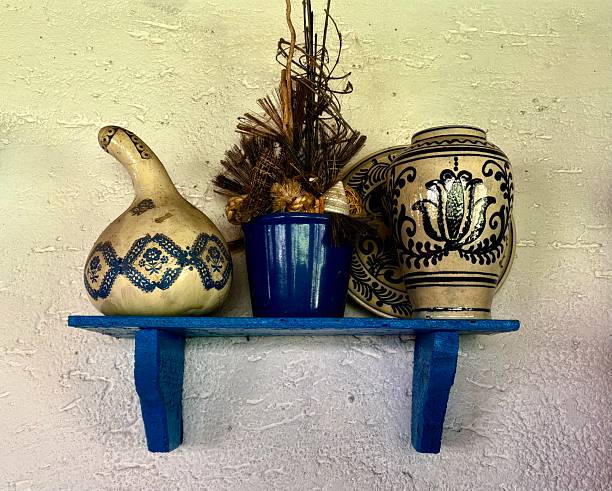 Traditional Serbian pottery and decorative items on a blue shelf. Handmade jugs and pots with characteristic patterns, along with a blue ceramic vase and decorative dried flowers, are displayed on a rustic blue wooden shelf attached to the wall with a rough, bright texture.