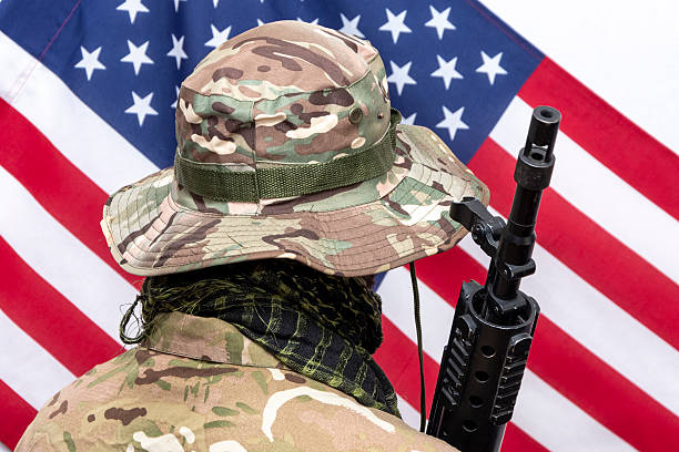 istock A soldier in camouflage uniform and panama hat with an automatic rifle on his shoulder against the background of the American flag., Concept: military operations, soldier of fortune, US Army. 2210770274