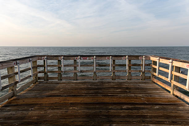 A weathered wooden fishing pier stretches over the ocean under a calm sky, with numbered stations for anglers visible along the railing.