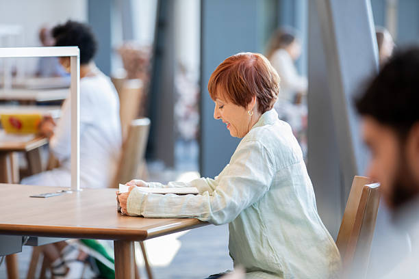 istock Senior woman sits at table in library and reads a book 2210784480