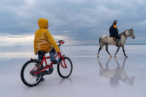 impressive encounter between a man on horseback and a boy riding a bicycle under rain clouds at the salt lake. A child wearing a yellow hooded jacket and a man wearing a hat. The child is watching the horseman riding a bicycle and loves the horse. An extraordinary photo emerged with the reflections on the water and the light leaking through the bulges. It was taken with a full-frame camera.