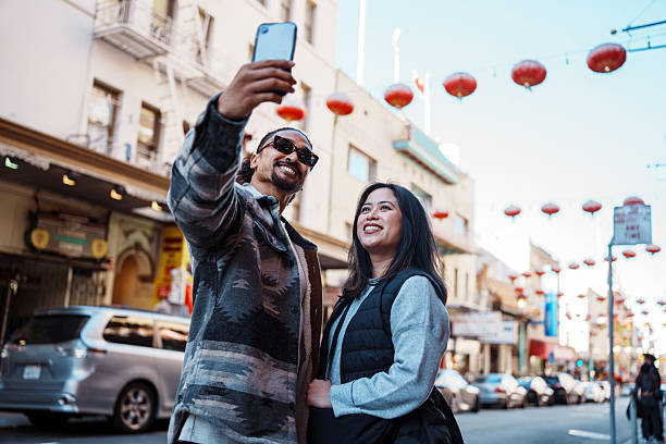 A Filipino couple in their 30s pose and take a selfie photograph with a smart phone while standing on a street in Chinatown with red Chinese Lanterns strung overhead during a fun weekend staycation exploring San Francisco, California, USA .