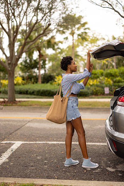 A young woman standing by a car, unloading groceries with a tote bag, surrounded by trees and greenery. Casual clothing and an outdoors setting highlight a relaxed and productive moment.