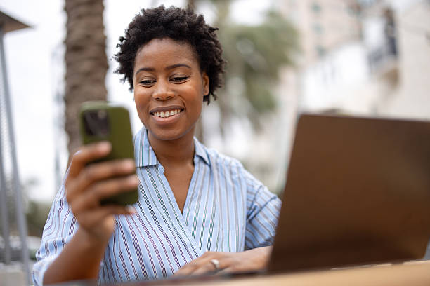 Smiling Woman Using Smartphone Outdoors With Laptop in Relaxed Setting