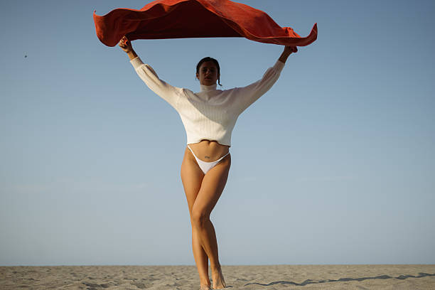 A woman stands gracefully on a sandy surface outside, holding red fabric against the sky. The expression and positioning emphasize freedom, harmony with nature, and a modern sense of elegance.
