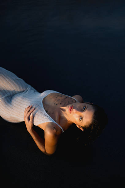 A serene woman in a white outfit lying on a sandy surface under dramatic sunset lighting.