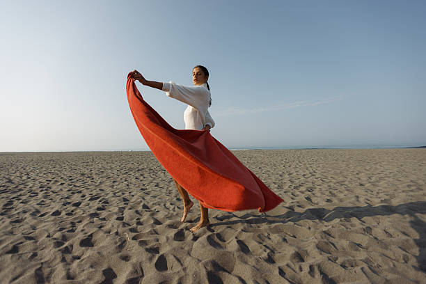 A graceful woman twirls on a sunlit beach, surrounded by serene sand and sea views, showcasing flowing fabric in autumn tones.
