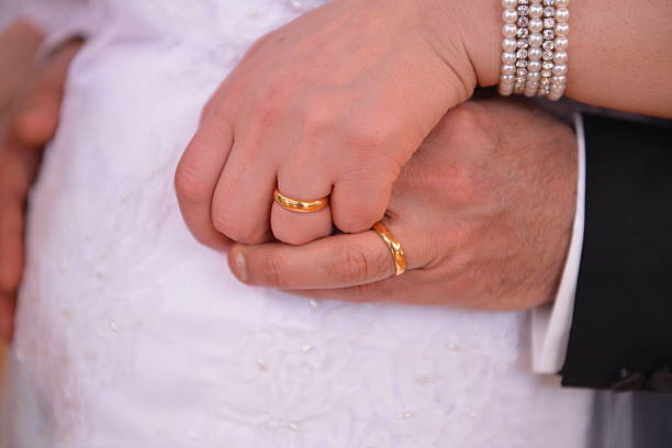 A close-up of the couple’s hands gently holding each other, their wedding rings gleaming softly. The bride's white dress forms the background while the groom’s dark suit adds contrast. Captured at waist level with soft ambient lighting to highlight the emotion and unity of the moment.
