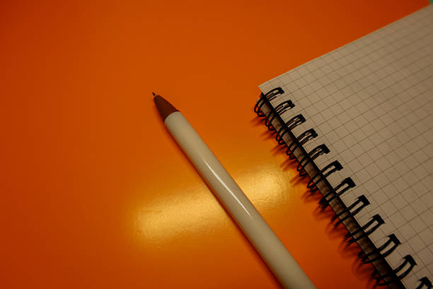 A white ballpoint pen with a brown tip lies next to an open grid notebook with a spiral binding on a bright orange background.
