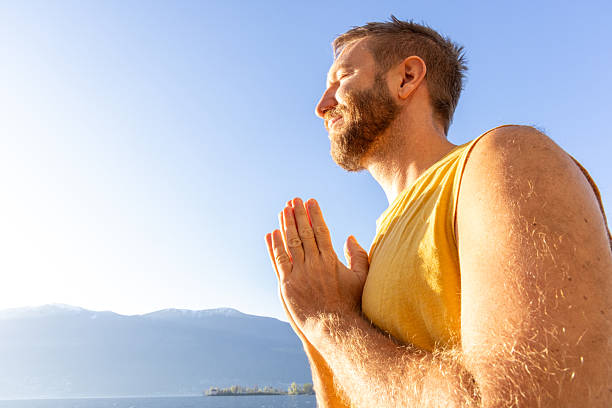 Man in a yellow tank top practicing meditation near a serene lake. Peaceful morning atmosphere with mountains in the background. Ideal for wellness and mindfulness themes.