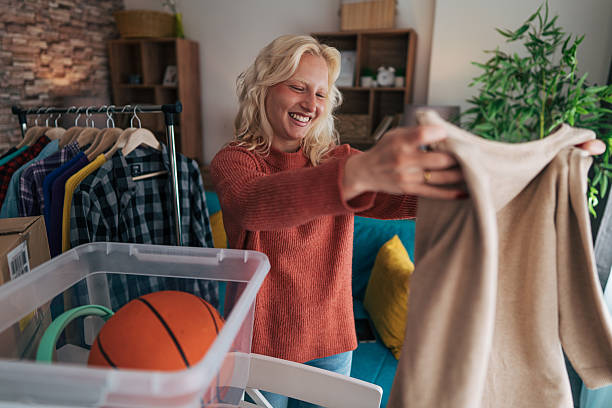 Young woman organizing clothes and items for donation or moving Young woman smiles while holding a sweater, organizing clothes on a rack and other items in a plastic box, preparing for donation, moving, or decluttering sorter machine stock pictures, royalty-free photos & images