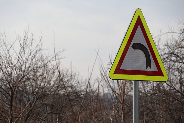 A cautionary warning sign is positioned to indicate a dangerous sharp curve on a winding road lined with bare trees