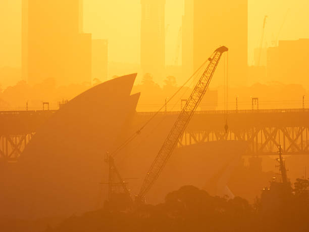 The sun is setting over the Sydney Opera House and Sydney Harbour Bridge.  In the foreground is a crane and communications mast of HMAS Brisbane at Garden Island naval base. In the background is the southern approach to the Sydney Harbour Bridge. This image was taken from Dudley Page Reserve, Dover Heights in the eastern suburbs of Sydney on a hot and sunny afternoon at sunset on 18 April 2025.