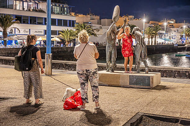 Arrecife, Lanzarote, Canary Islands, Spain - March 11th 2025: This lively nighttime image captures three middle-aged female tourists playfully interacting with a statue in Arrecife, Lanzarote. Their joyful expressions and animated gestures bring a sense of spontaneity to the scene, highlighting the charm of public art in urban spaces. The illuminated waterfront in the background adds depth to the composition, while the statue itself stands as a cultural landmark, inviting engagement and curiosity.
