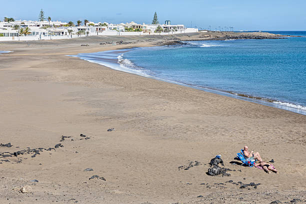 Arrecife, Lanzarote, Canary Islands, Spain - March 14th 2025:  This peaceful image captures a couple enjoying the solitude of an empty beach just outside Arrecife, Lanzarote. Seated on beach chairs near the water’s edge, they embrace the tranquility of the Atlantic, with soft waves rolling onto the shore. The golden sand stretches undisturbed, while the distant white buildings and palm trees hint at the island’s coastal charm. The clear sky and calm ocean enhance the serene atmosphere, making this scene a perfect representation of quiet retreat and relaxation.