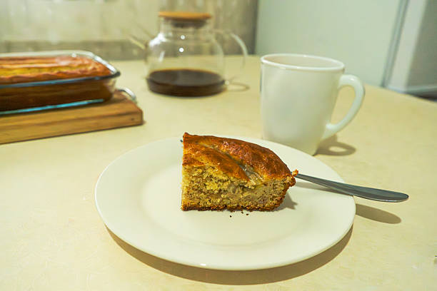 A cozy scene featuring a slice of homemade banana bread on a white plate with a spoon, paired with a hot cup of tea. A glass dish filled with more banana bread and a clear teapot filled with brewed tea sit in the background. Set on a clean light-colored surface, this warm and inviting image captures a peaceful tea-time moment—perfect for food blogs, recipe websites, or lifestyle content that highlights homemade comfort and simple pleasures.