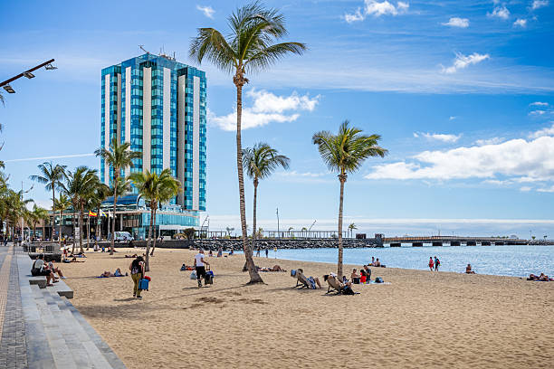 Arrecife, Lanzarote, Canary Islands, Spain - February 27th 2025:  This image showcases the Gran Hotel in Arrecife, Lanzarote, standing tall against the backdrop of a palm-fringed beach. The foreground features golden sands dotted with lush palm trees, creating a tropical contrast to the modern architecture of the island’s tallest building. The deep blue Atlantic stretches beyond, while the clear sky and gentle waves enhance the serene yet vibrant atmosphere of this coastal retreat. Ideal for themes related to travel, luxury destinations, and the harmony between nature and urban landscapes.