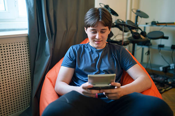 A 14-year-old teenager is captured in a quiet moment of focus, playing with a vintage handheld game console—such as a Game Boy, PSP, or another early-generation device. The teen holds the console with both hands, eyes locked on the small screen, completely absorbed in gameplay. This image blends youthful energy with retro charm, evoking themes of analog entertainment, nostalgia, and the timeless appeal of gaming across generations.