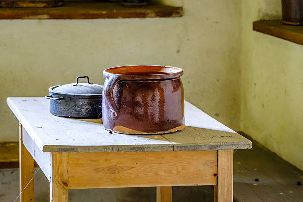 A ceramic cooking pot and a cast iron skillet rest on a wooden table in a rustic kitchen, with warm natural light illuminating the scene, creating a cozy atmosphere.