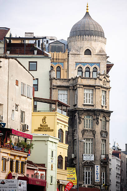 Istanbul, Turkey - November 21, 2015: A historic building with a dome at the top is seen alongside multi-story structures. The facade features arched windows, columns, and balconies. Various business signs are visible at street level. Buildings with different architectural styles appear on both sides.