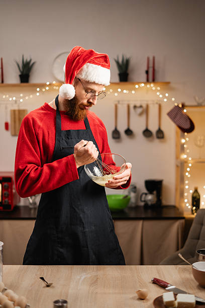 istock Baker in Santa hat whisking eggs in a glass bowl, preparing ingredients for baking in his kitchen. Concept of home baking and culinary preparation. 2210902122