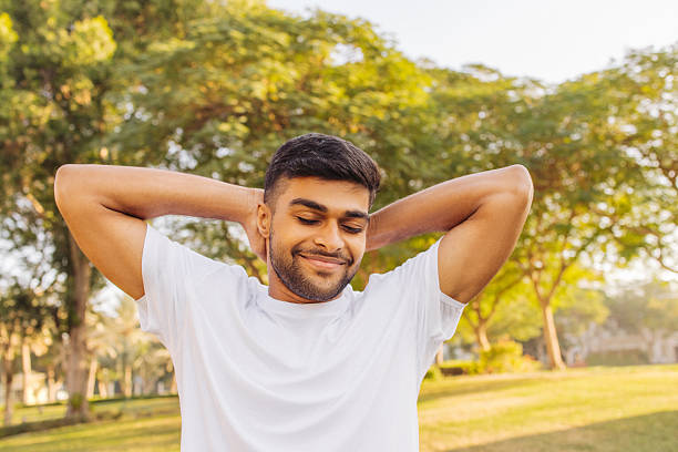 A young man enjoying a serene moment in a sunlit park, exuding a sense of joy and relaxation. He is wearing a white shirt, smiling with eyes closed, and has his hands placed calmly behind his head. The trees and greenery in the background add to the peaceful and cheerful ambiance.