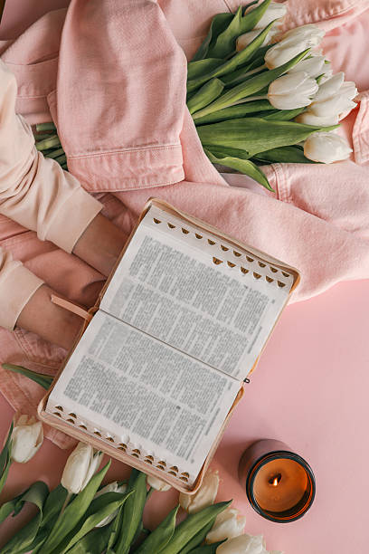 istock Open Bible held in hands over a pink background, surrounded by white tulips, a lit candle, and a pink denim jacket, creating a peaceful devotional setting 2210918422