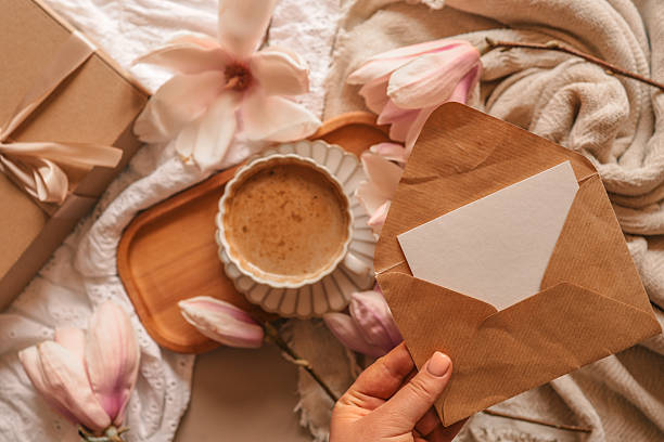 istock A flat lay with a kraft envelope and blank card in hand, next to a wrapped gift box, a coffee cup, blooming magnolia flowers, a lit candle, and soft cozy fabrics 2210918523