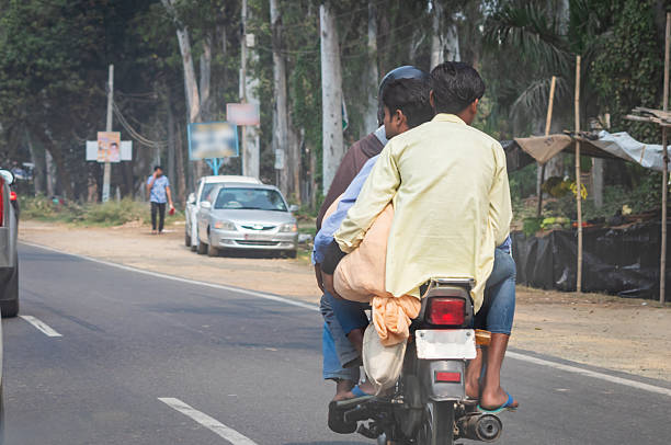 An Indian man in a black riding jacket, seated on a motorcycle, looks with a smiling and confident expression. Set against a lush green backdrop on a rural road with another biker passing in the background, the scene evokes a sense of freedom, tranquility, and the open road.