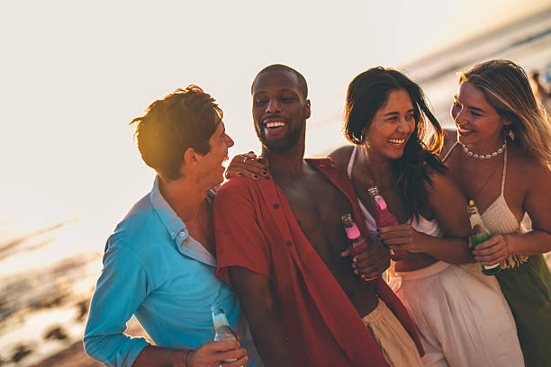 Four diverse friends gathered by the beach at sunset, sharing drinks and enjoying each other's company in a lively, joyful atmosphere. A celebration of friendship and summer coastal moments.