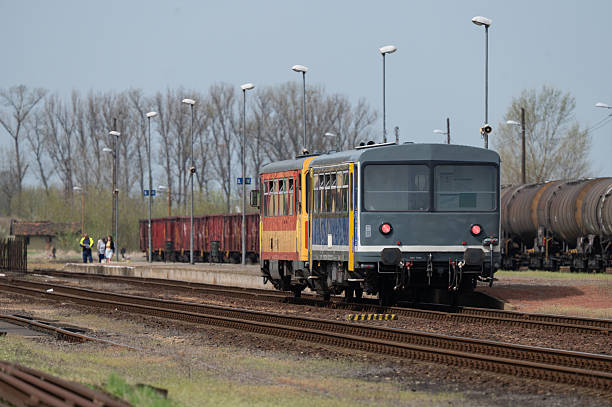 A small, vintage passenger train in vibrant colors stands at a quiet rural railway station surrounded by freight cars and fuel tanks. The background features leafless trees and overcast lighting, creating a nostalgic, slightly melancholic mood. The scene evokes a sense of stillness and the charm of old rail travel.