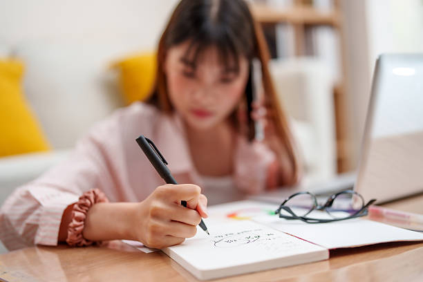 istock young woman is working from home, focused on her notes while holding phone. She writes with pen on notebook, surrounded by laptop and glasses, creating productive atmosphere.freeelance 2210962343