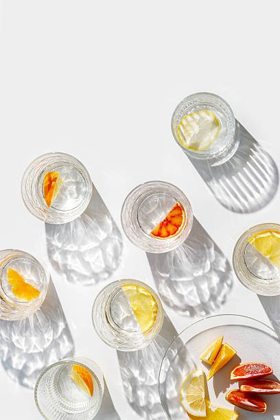 istock Crystal glasses with citrus slices in water on white background with reflections at sunlight, modern, minimal composition with clear glass cups with water, citrus fruit as lemon, orange on plate 2210967739